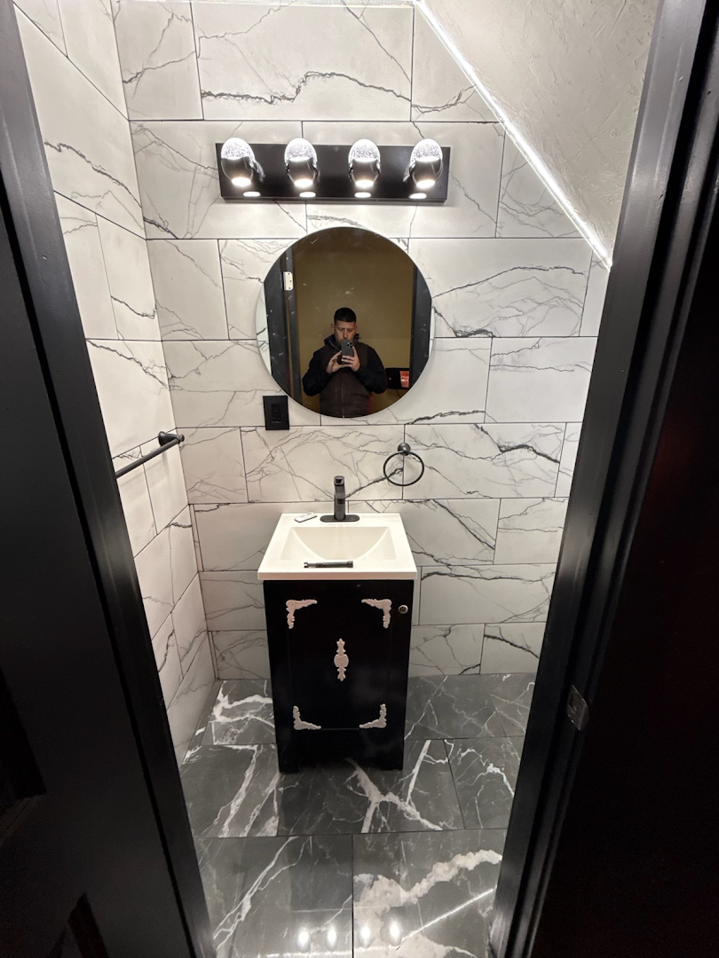 Powder room with marble tile walls, four-light vanity bar, round black mirror and black vanity
